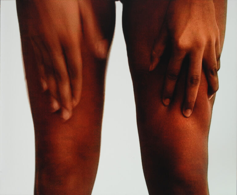 A set of medium skin-toned legs are visible from the upper thigh to just below the knee. Hands reach down from the top of the frame and rest on the skin above the knee.