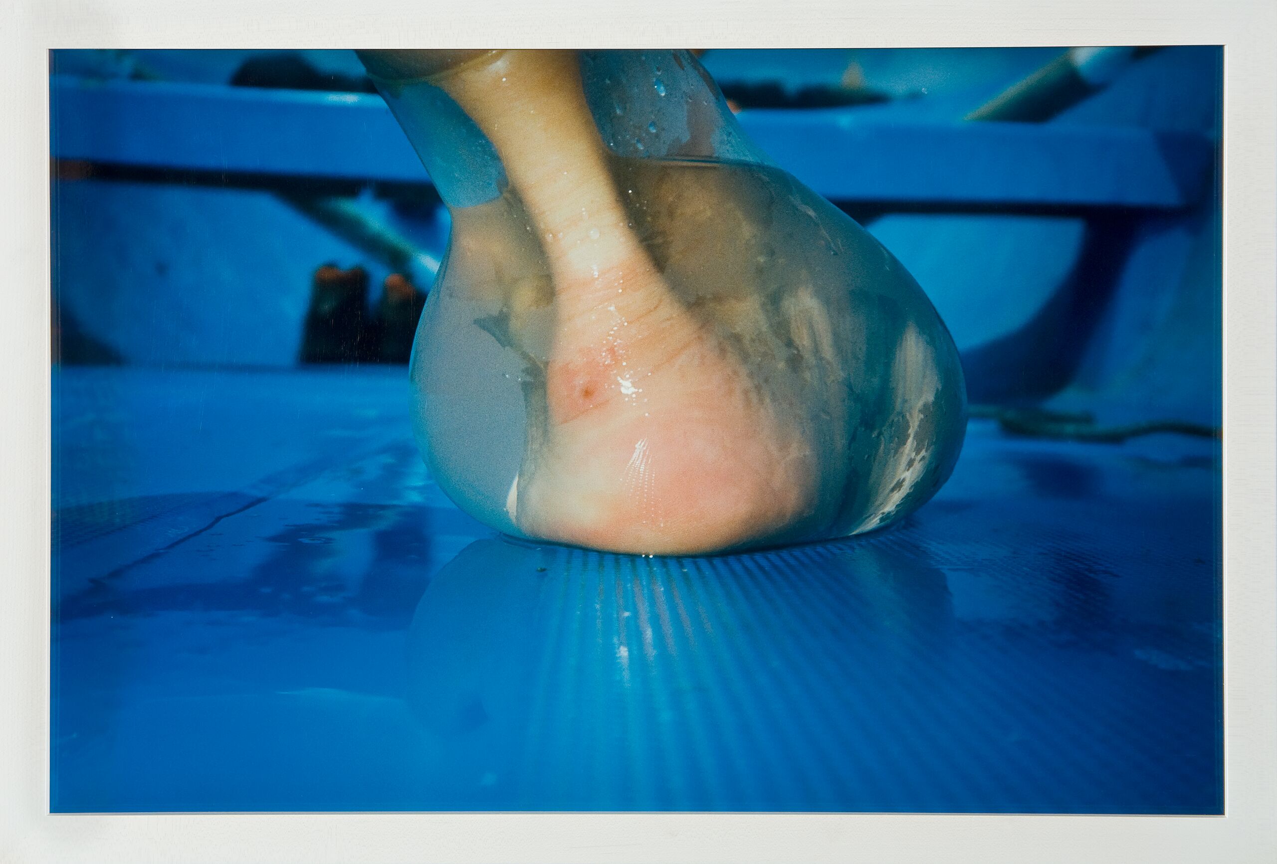 A photograph shot at ground level captures an extreme close-up of a heel and ankle from behind. Enclosed in a water-filled latex balloon that distorts its shape, the foot rests upon a wet, sky blue surface that resembles the interior hull of a rowboat.