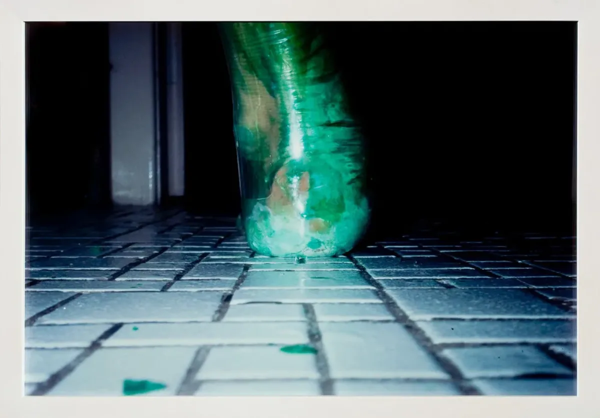 A photograph shot at ground level captures an extreme close-up of a heel and ankle from behind. Enclosed in a clear latex balloon filled with viscous green liquid that distorts its shape, the foot rests upon a floor of tiny, square, white tiles, like those used in bathrooms.