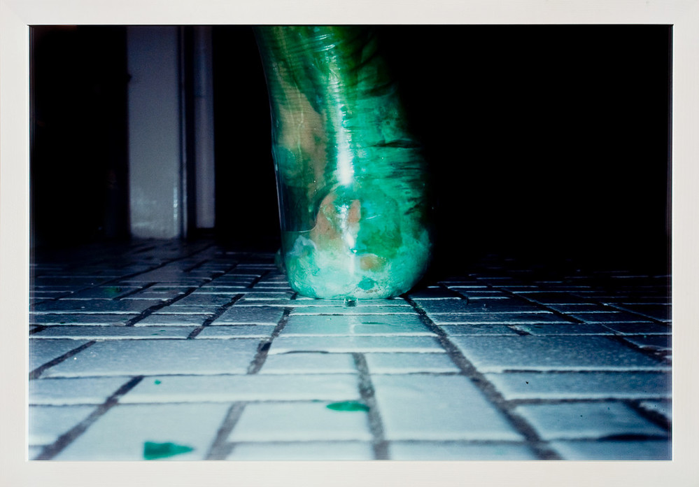 A photograph shot at ground level captures an extreme close-up of a heel and ankle from behind. Enclosed in a clear latex balloon filled with viscous green liquid that distorts its shape, the foot rests upon a floor of tiny, square, white tiles, like those used in bathrooms.