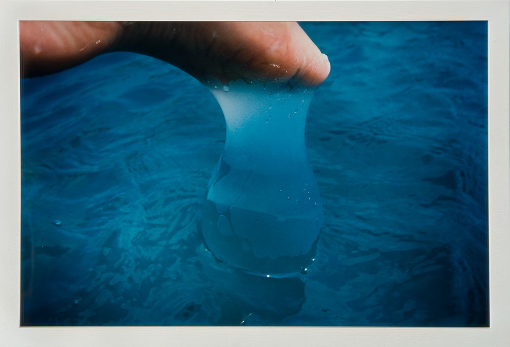 The big toe, arch, and lower heel of a left foot appear beneath the upper frame of a photograph. A water-filled latex balloon slips off the foot, forming a tear shape that dangles from the toe and connects with the rippling surface of blue water that dominates the composition.