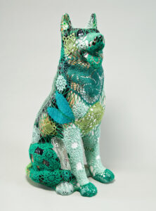 A mass-produced, ceramic German shepherd sits upright and alert with mouth open and tongue extended as if panting. Panels of elaborate hand-crafted crochet in shades of mint, citron, and emerald green form a skin-tight web that entirely envelops the dog.