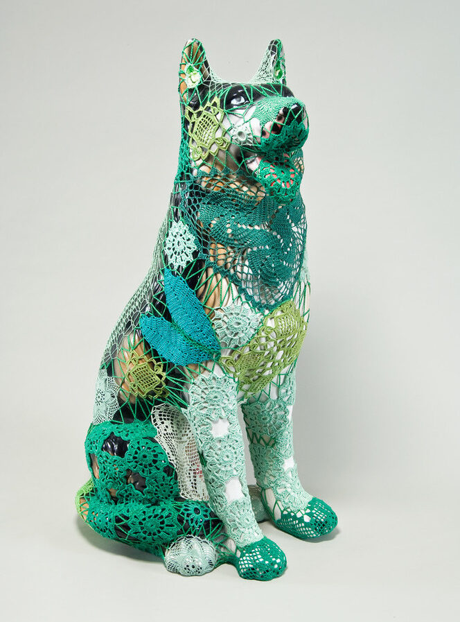 A mass-produced, ceramic German shepherd sits upright and alert with mouth open and tongue extended as if panting. Panels of elaborate hand-crafted crochet in shades of mint, citron, and emerald green form a skin-tight web that entirely envelops the dog.