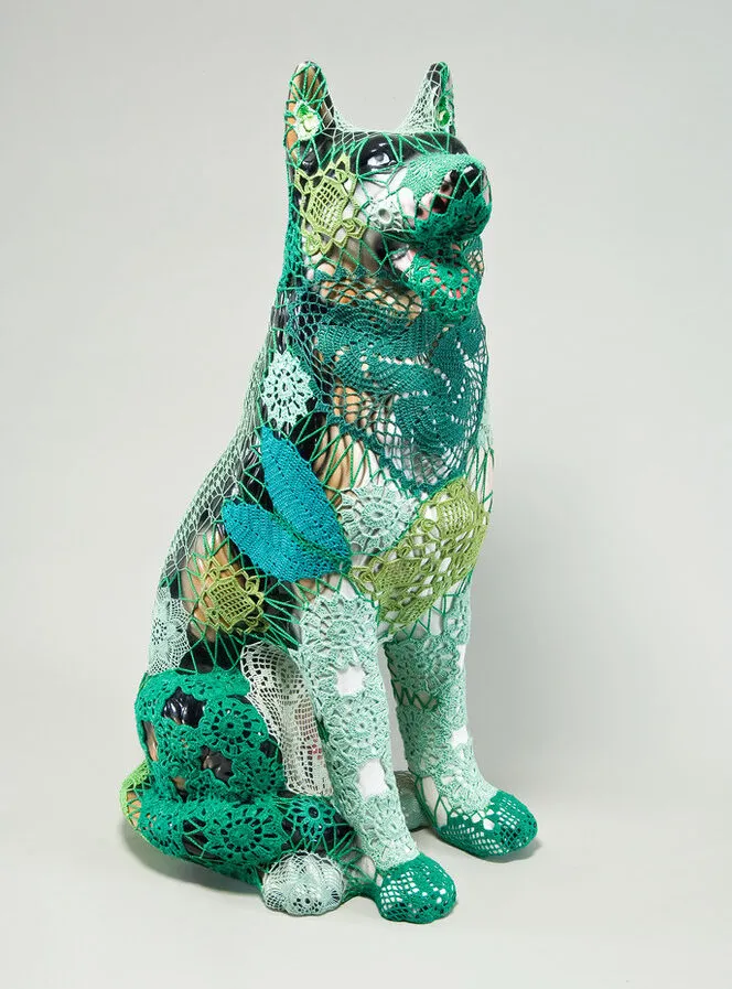 A mass-produced, ceramic German shepherd sits upright and alert with mouth open and tongue extended as if panting. Panels of elaborate hand-crafted crochet in shades of mint, citron, and emerald green form a skin-tight web that entirely envelops the dog.