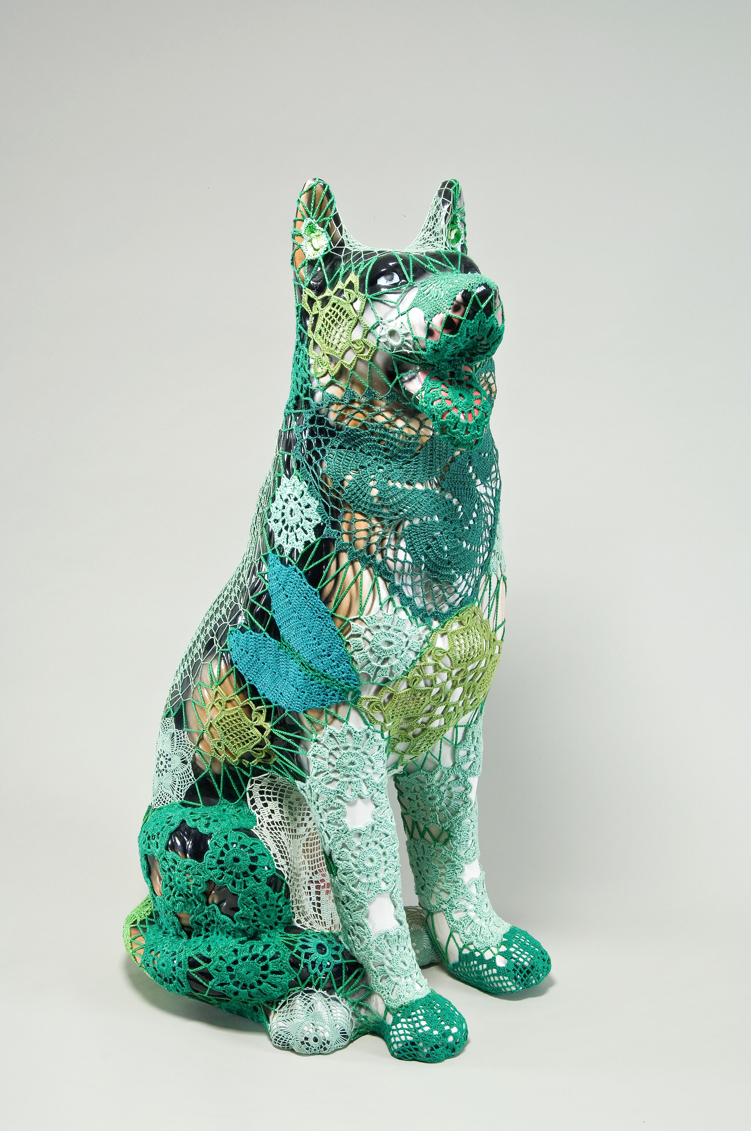 A mass-produced, ceramic German shepherd sits upright and alert with mouth open and tongue extended as if panting. Panels of elaborate hand-crafted crochet in shades of mint, citron, and emerald green form a skin-tight web that entirely envelops the dog.
