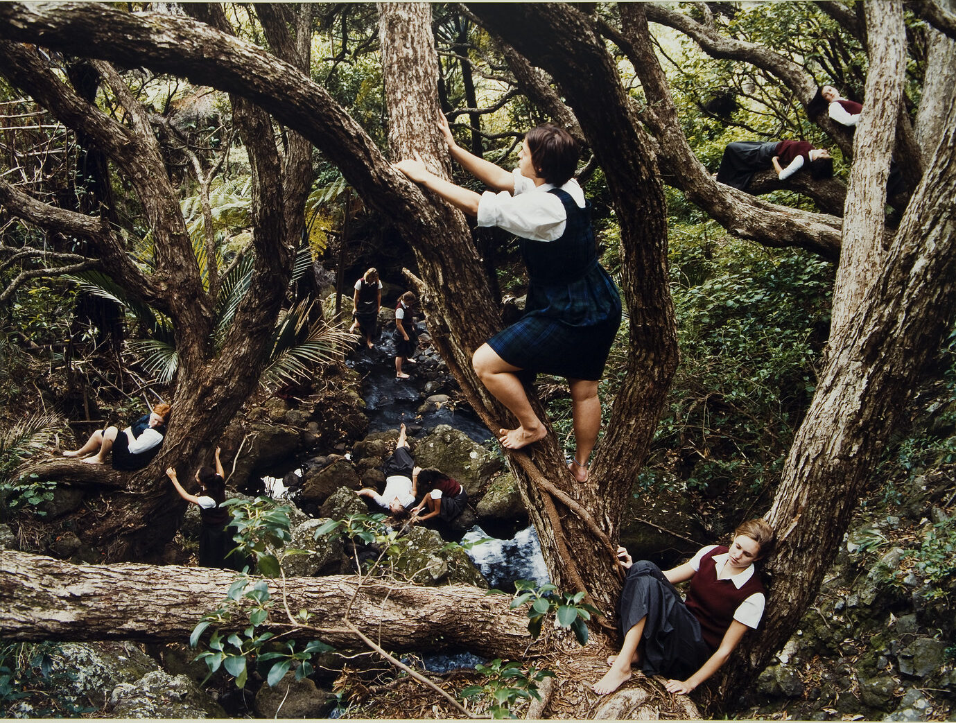 Color photograph of a dense forest with a light skin-toned girl with bare feet climbing a tree at center. Below her to the right is another light skinned girl sitting against the trunk of a tree. Other figures are scattered throughout the forest in the background.