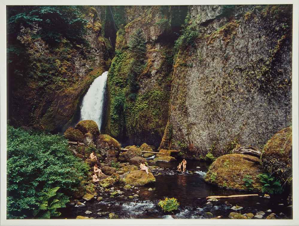 Photograph of a splendid landscape scene featuring a cliff, waterfall and stream. Nude mothers with their children sit on rocks or play in the stream. The human figures are dwarfed by blocks of green foliage in the lower left corner and walls of steep rock covered subtly by moss.