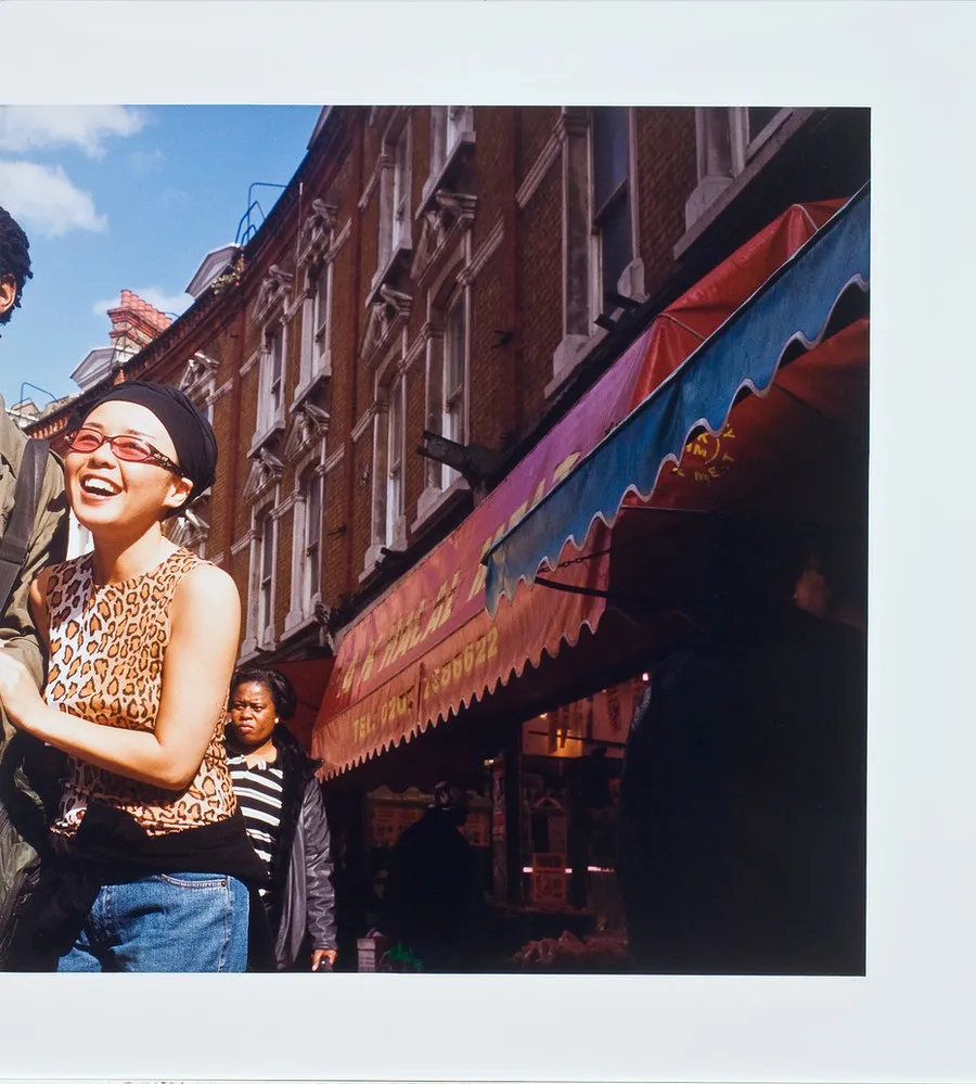 Color photograph of a street scene. Red brick buildings with colorful awnings are on the right and a smiling woman on the left. She holds the arm of a companion who has been cropped out of the left side of the image.