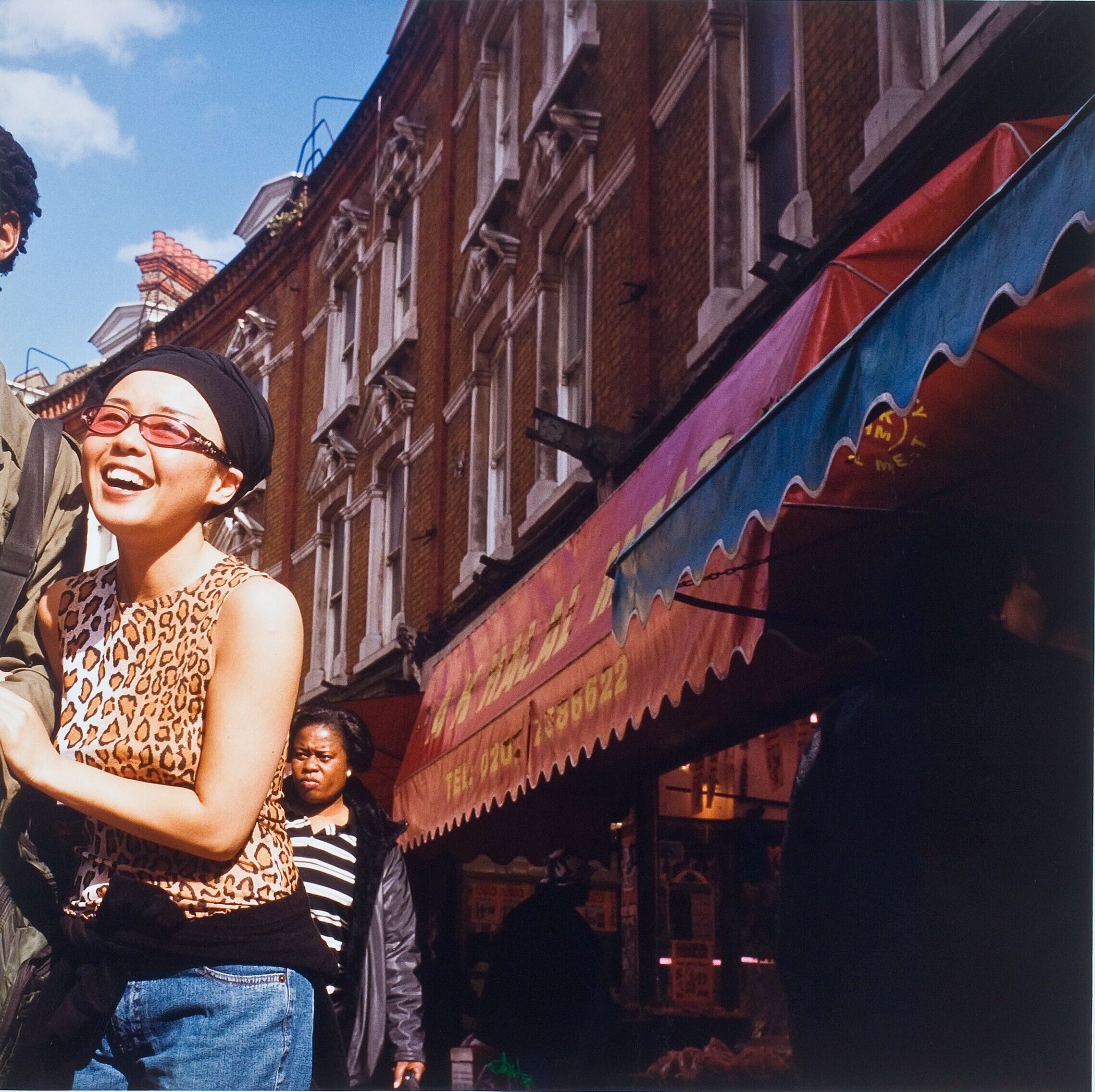 Color photograph of a street scene. Red brick buildings with colorful awnings are on the right and a smiling woman on the left. She holds the arm of a companion who has been cropped out of the left side of the image.