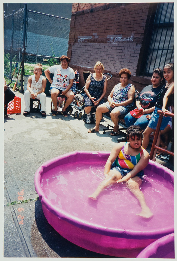 Color photograph of a group of figures with medium skin tone in an urban setting. Six figures sit on chairs in semi-circle and one young woman sits in a plastic swimming pool in the foreground. One more figure is almost completely cropped out of the image at the upper left.