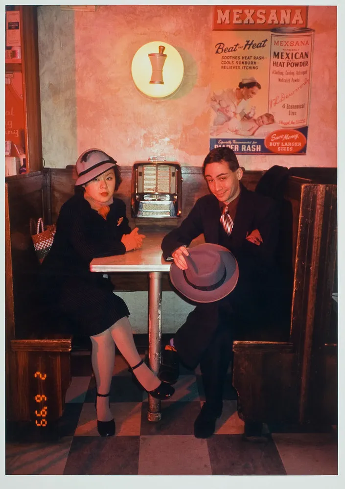 Color photograph of a man and woman sitting at a diner booth, looking at viewer. Woman on left wears gray hat with black trim. Man on right wears a dark suit and shiny silver tie. He holds his gray hat in his hands. Behind them on the table is a small juke box.