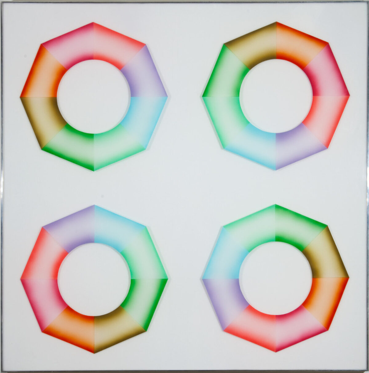 Four hard-edged octagonal rings, divided into wedges of brown, orange, pink, red, purple, blue, mint green, and grass green, sit on a square, white surface. Dark at the inner and outer edges and light in the center, the colors create the illusion of curved 3-dimensional objects.