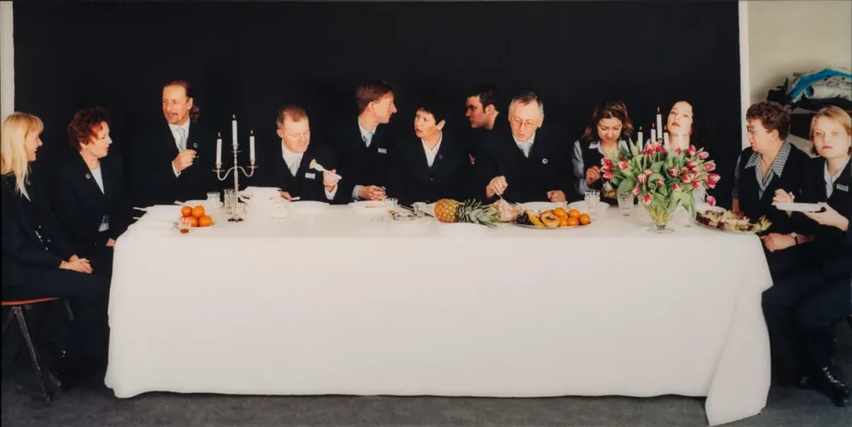 Twelve light-skinned men and women in dark suits sit on the ends and far side of rectangular table situated horizontally to the viewer. Covered with a white cloth, the table holds 2 candelabra, a flower bouquet, and assorted fruit. The people eat, chat, or look out at viewers.