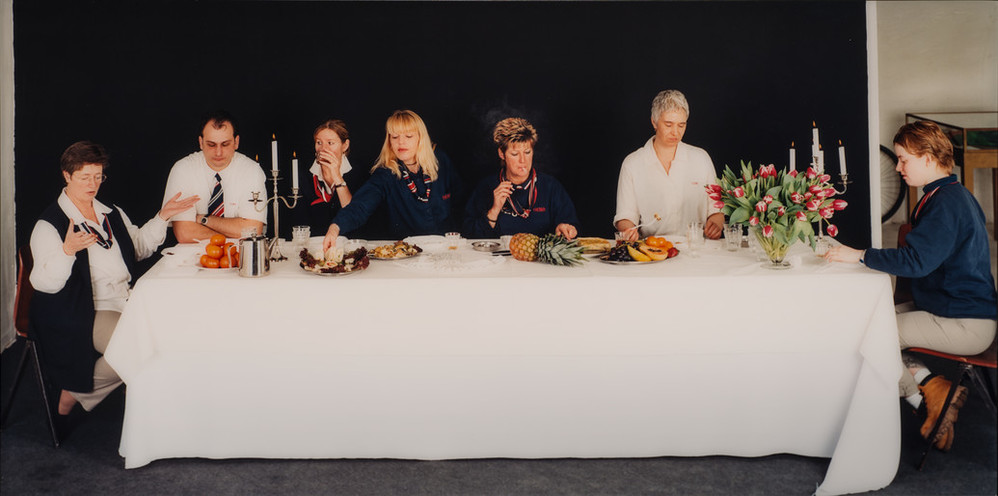 In front of a black background, seven light-skinned, plainly dressed women and men sit around a rectangular table. Food, candles and tulips sit on the white table cloth. The women and men gesture, smoke, drink, and eat, but do not engage with each other or the viewer.
