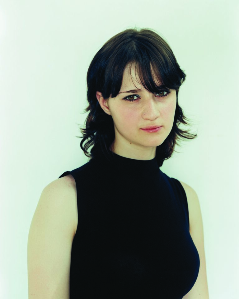 Paired photographic portraits capture a young, light-skinned woman at different life moments. At left, she wears a black sweater and gazes out shyly from beneath her dark bangs. At right, clad in an olive drab, military shirt, she makes eye contact with viewers more confidently.