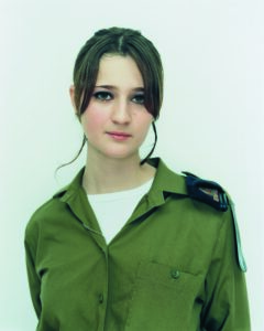 Paired photographic portraits capture a young, light-skinned woman at different life moments. At left, she wears a black sweater and gazes out shyly from beneath her dark bangs. At right, clad in an olive drab, military shirt, she makes eye contact with viewers more confidently.