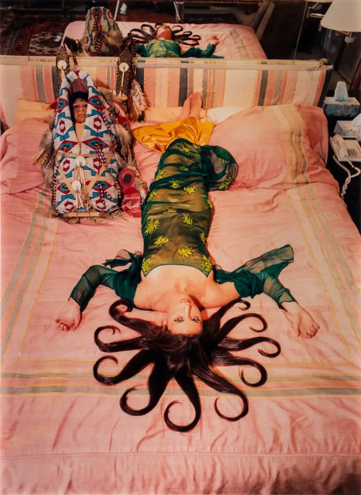 A slim, light-skinned woman wearing a long, emerald-green dress gazes up at the viewer from the foot of the bed, her bare feet near the headboard. Brown hair radiates from her head in 10 curled, snake-like segments. Her arms, bent at the elbows, extend from her body to form a W.