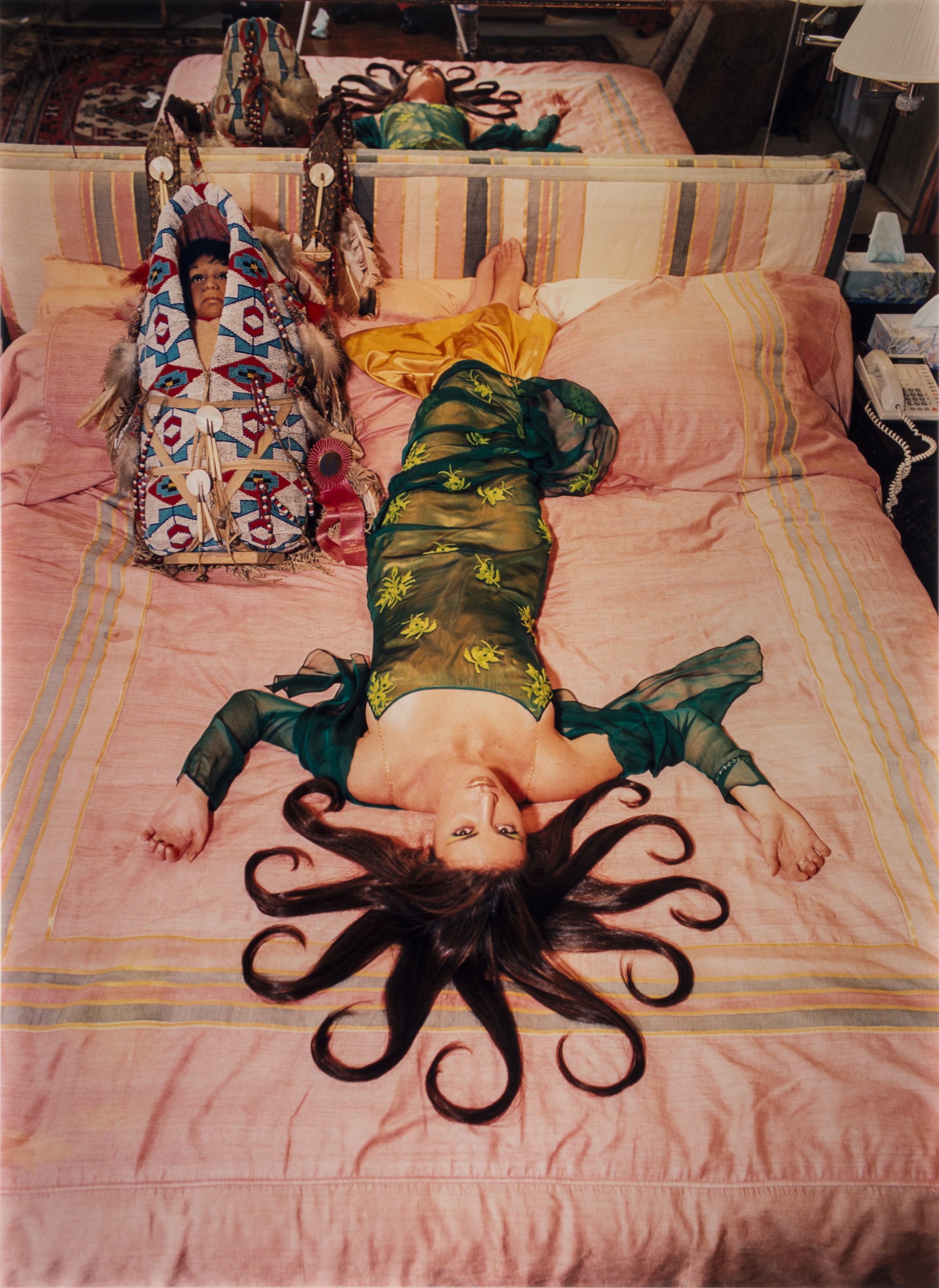 A slim, light-skinned woman wearing a long, emerald-green dress gazes up at the viewer from the foot of the bed, her bare feet near the headboard. Brown hair radiates from her head in 10 curled, snake-like segments. Her arms, bent at the elbows, extend from her body to form a "W."