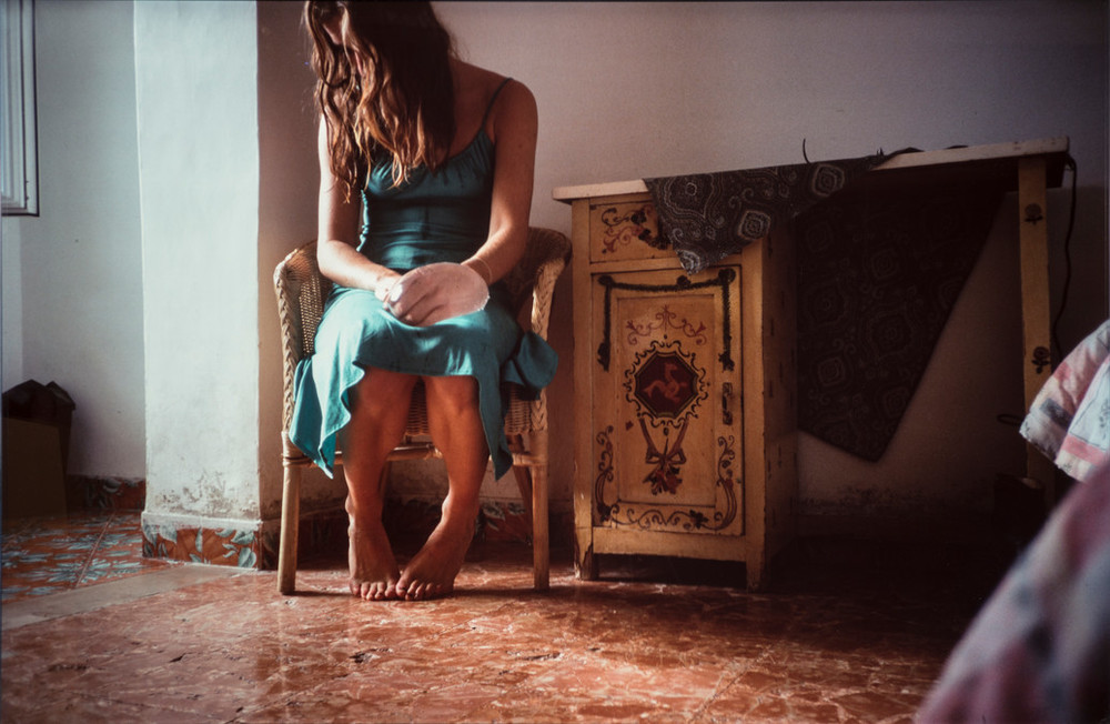 A photograph portrays a woman clad in an aqua blue slip dress. She sits in a wicker chair in a naturally lit room with red marble flooring. Her head and shoulders slump forward so that her long brown hair obscures her face. A water-filled latex balloon encloses her left hand.