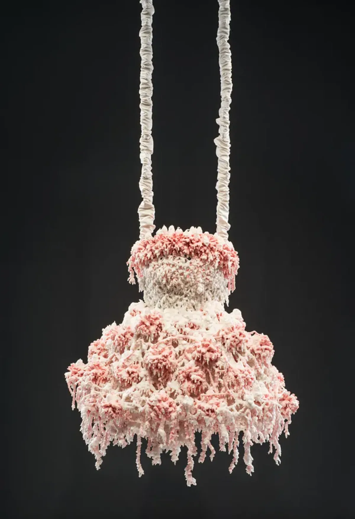 Myriad layers of melted pink and white wax encrust and obscure the metal armature for this abstract sculpture, which hangs from satin-wrapped chains. Its color and shape, as well as the bumpy, lacy texture, evoke a frilly tutu, lavishly frosted wedding cake, or coral accretions.