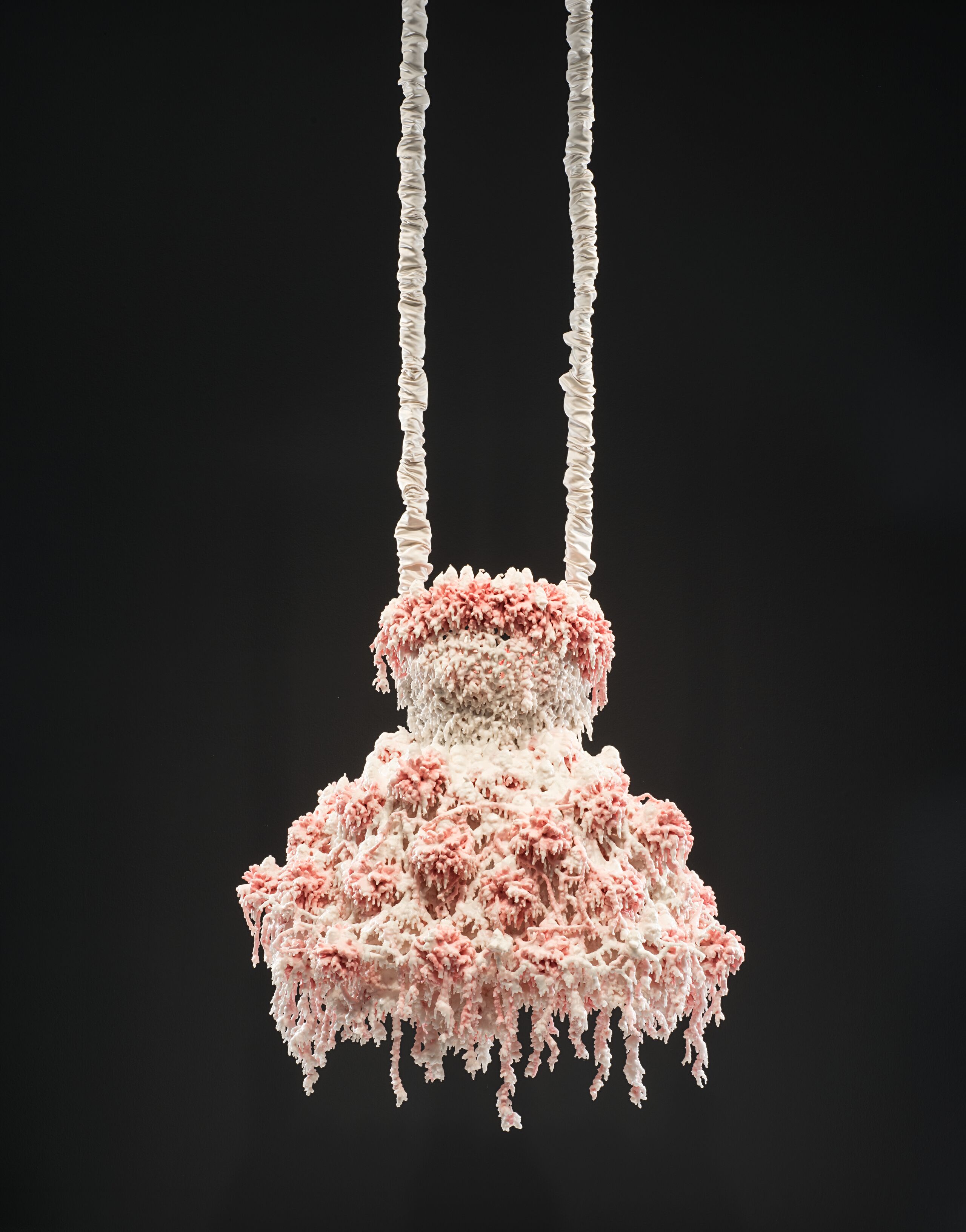 Myriad layers of melted pink and white wax encrust and obscure the metal armature for this abstract sculpture, which hangs from satin-wrapped chains. Its color and shape, as well as the bumpy, lacy texture, evoke a frilly tutu, lavishly frosted wedding cake, or coral accretions.