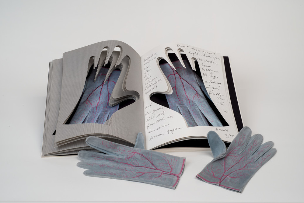 An open book into whose pages the shapes of hands have been cut. Gray gloves painted with red veins are visible through the cutouts. An identical pair of gloves rest next to the book.