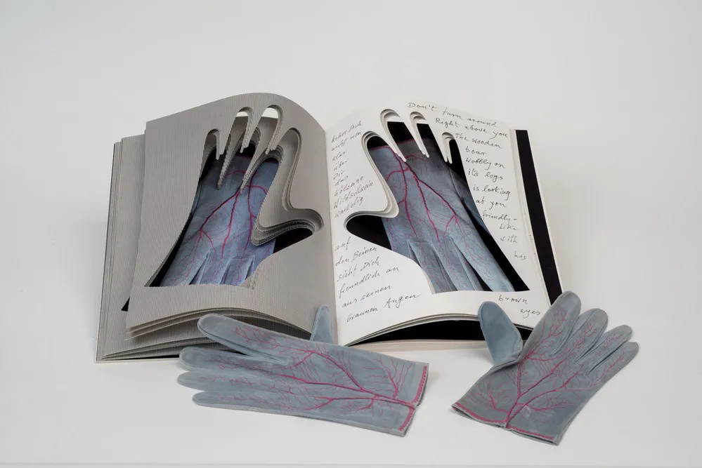 An open book into whose pages the shapes of hands have been cut. Gray gloves painted with red veins are visible through the cutouts. An identical pair of gloves rest next to the book.