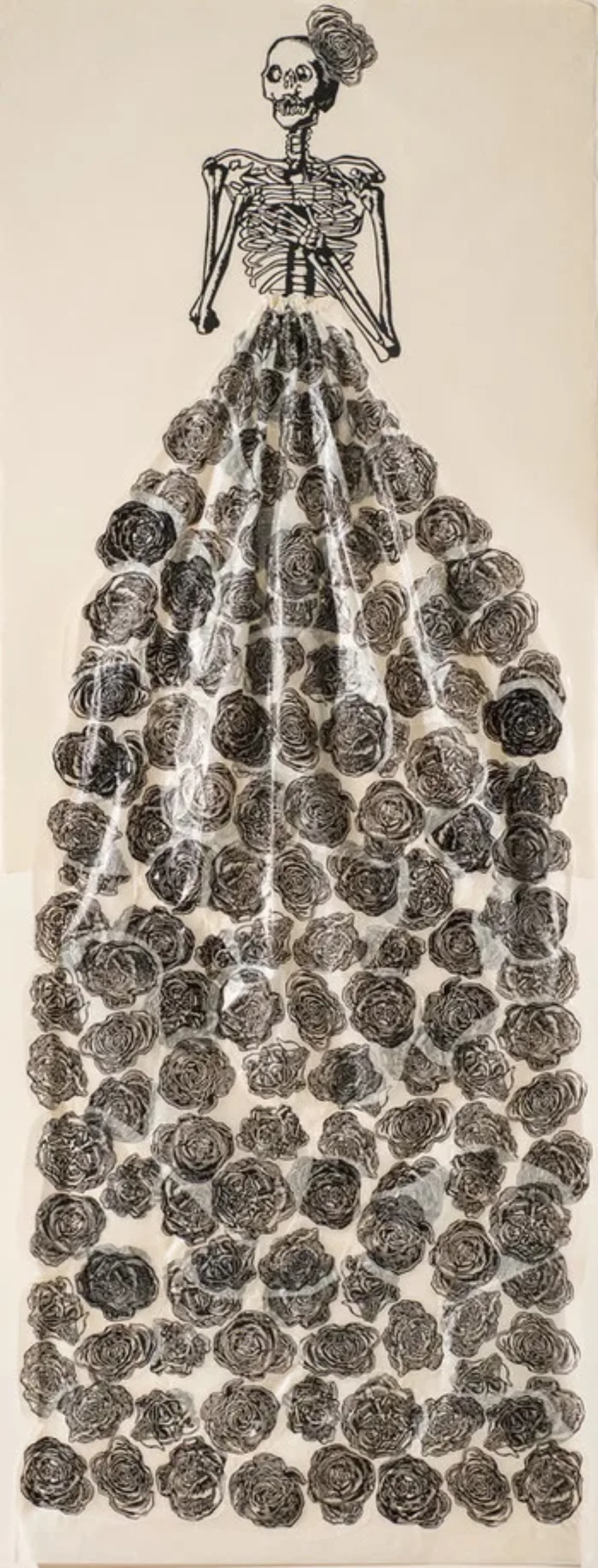 A very tall and narrow, black-and-white print pictures a skeleton with folded arms, a rose fascinator perched on its head. The figure wears a collaged, three-dimensional, billowing skirt made of tissue and printed with roses.