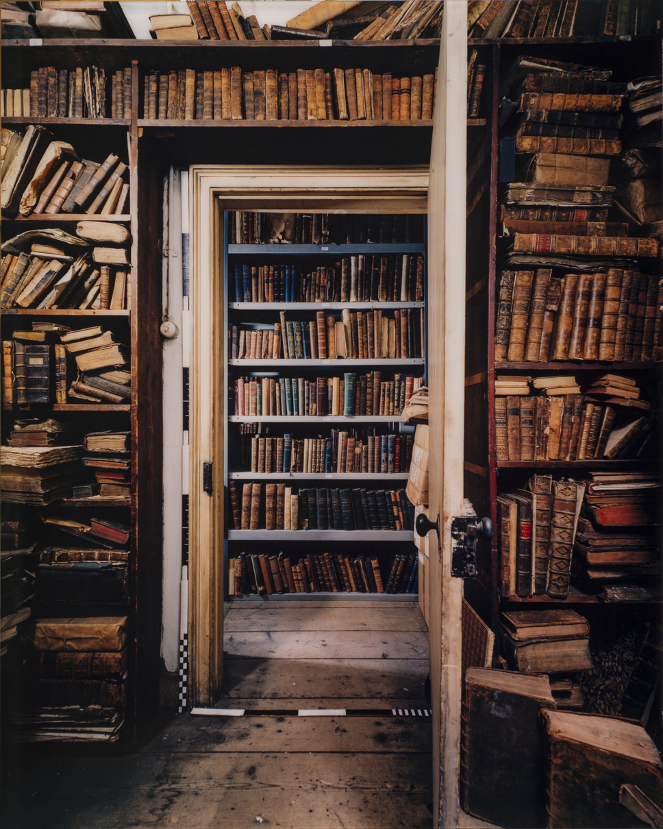 Color photograph of room with old books on bookshelves that stretch from floor to ceiling. In the center is an open door revealing another room with floor to ceiling shelves filled with books.