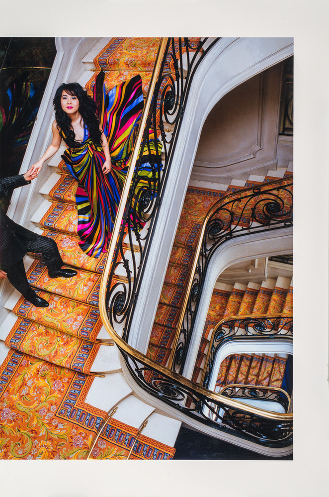 A bird's eye view of a woman with long, dark hair in a brightly-colored gown descending an ornate spiral staircase of white marble and colorfully patterned rug. Her right hand is being held by a figure in a black suit whose head and shoulders are cropped out of the image.