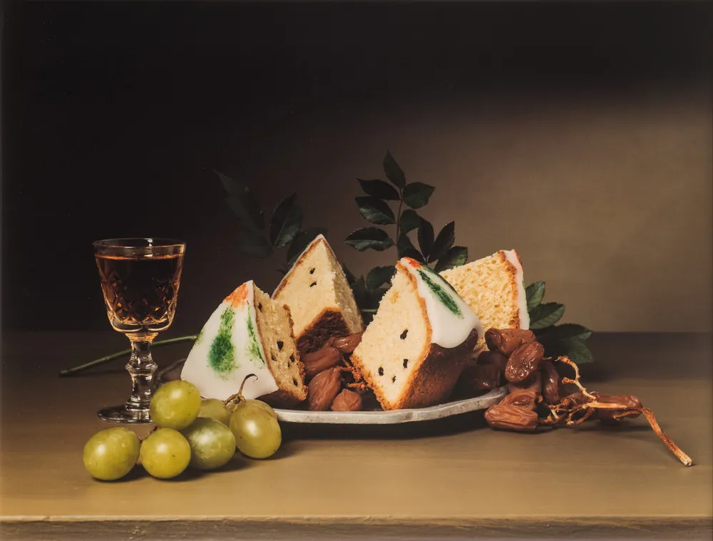 A full crystal sherry glass and plate mounded with raisins and iced cakes occupy the middle ground of a still-life photograph. Luscious green grapes sit in the left foreground, and greenery rises in the background. The image explicitly imitates18th-century, still-life paintings.