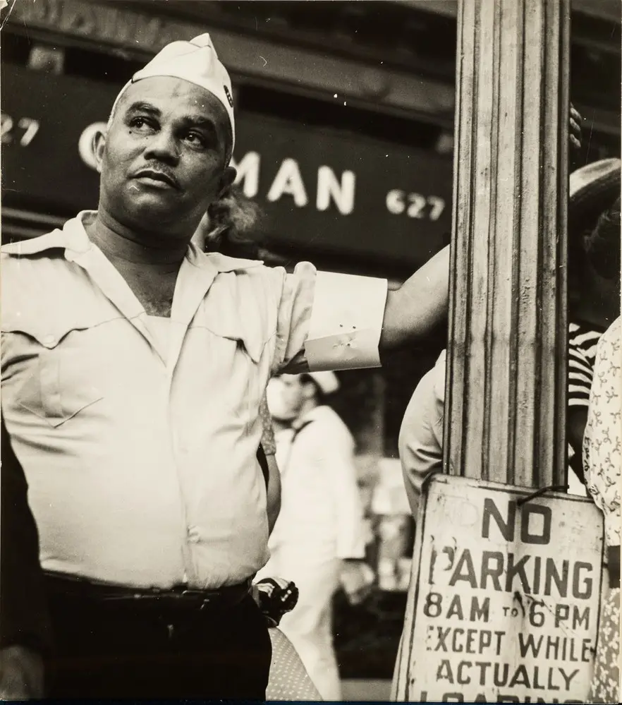 Candid street-scene photograph of a dark-skinned man wearing a WWII era military uniform. He gazes up and the his left, his left arm raised as he casually holds on to a lamp post with a "No Parking" sign attached to it.