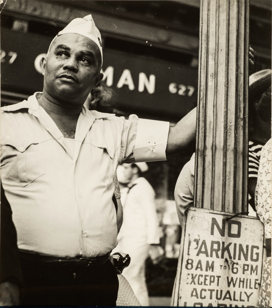 Candid street-scene photograph of a dark-skinned man wearing a WWII era military uniform. He gazes up and the his left, his left arm raised as he casually holds on to a lamp post with a 