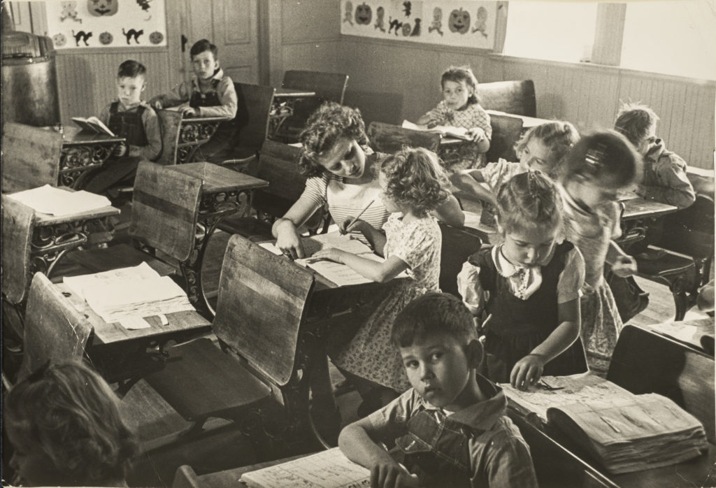 Black and white photograph of a primary school classroom in 1948. Light-skinned children sit at dark wood desks attending to books and papers or looking directly into the camera. At the center, the young teacher assists a girl with her lesson.