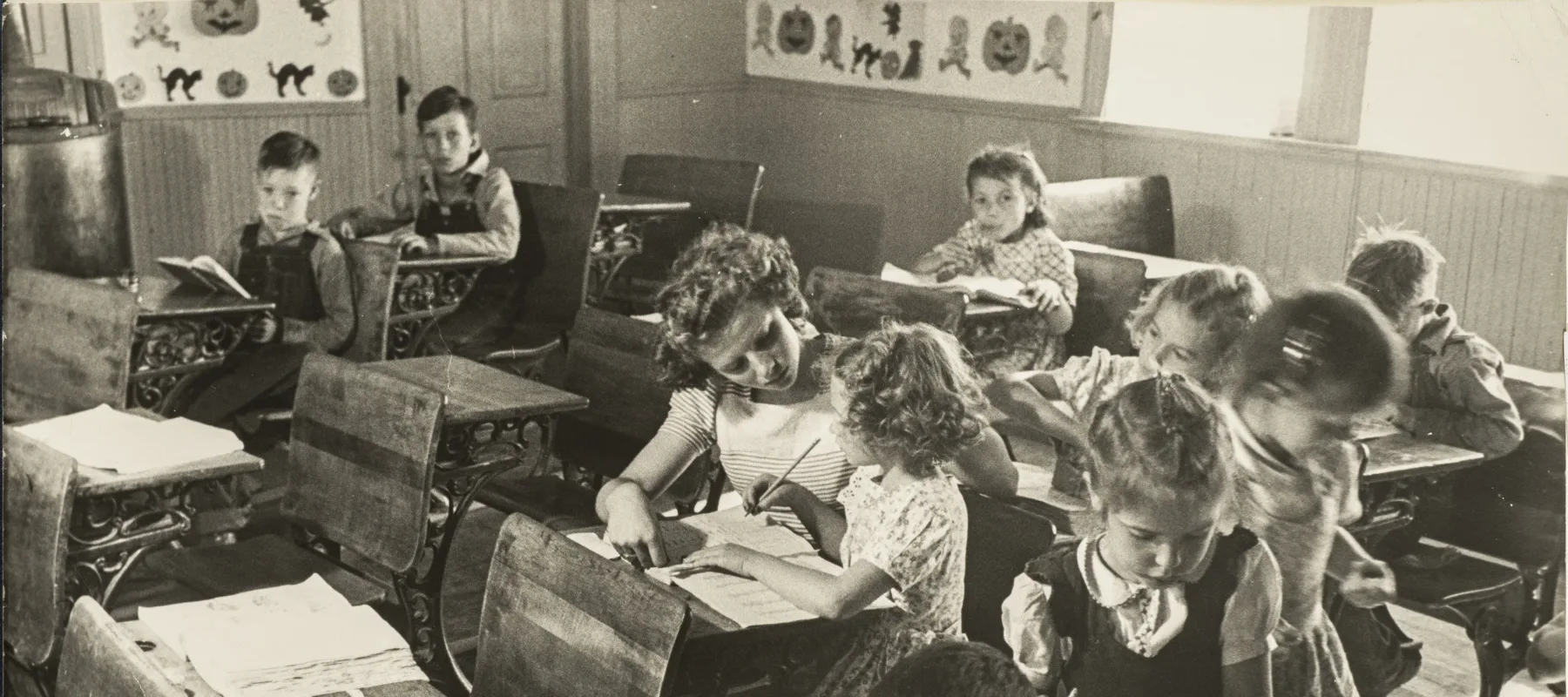 Black and white photograph of a primary school classroom in 1948. Light-skinned children sit at dark wood desks attending to books and papers or looking directly into the camera. At the center, the young teacher assists a girl with her lesson.