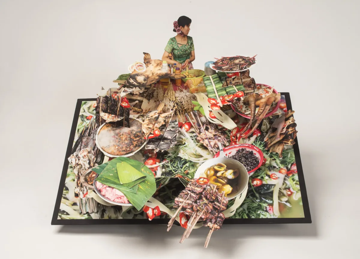 Large-scale pop-up book with a dark haired woman behind a huge display of food. She wears a green dress and purple flowers in her hair and turns to look over her left shoulder. The food explodes forward with bowls of soup, skewered meat, banana leaves, pig tail, and more.