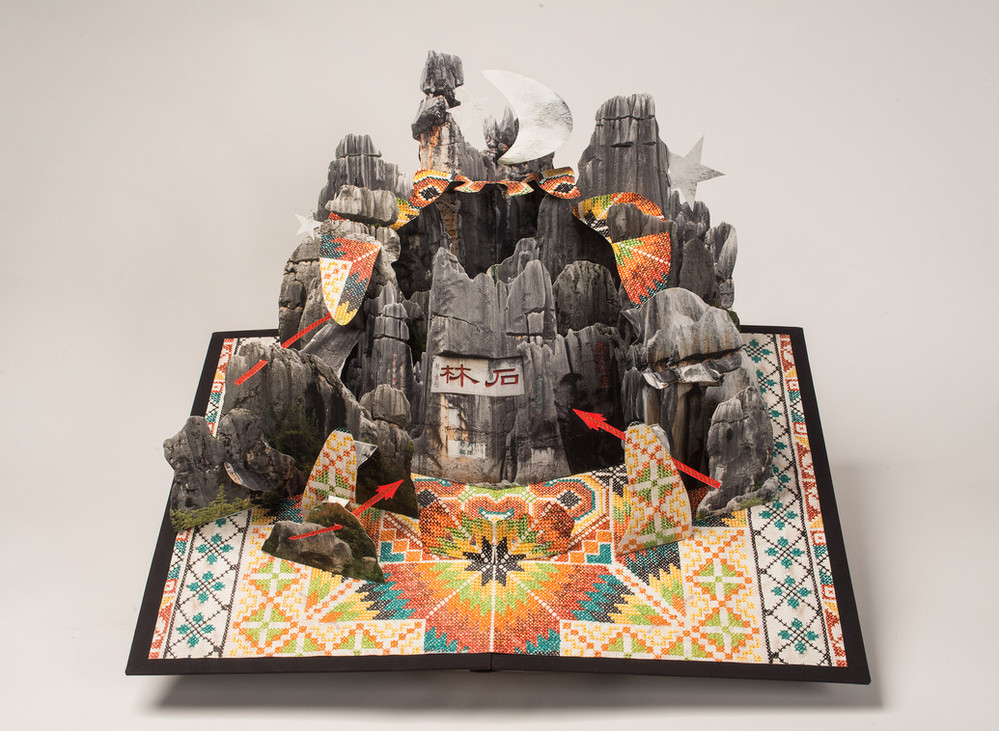 Large-scale pop-up book with a group of limestone formations resembling weathered tree trunks sits on top of a hand-embroidered base of colorful geometric patterns that repeat in abstract shapes over the center of the piece. A crescent moon and single star peek from behind.