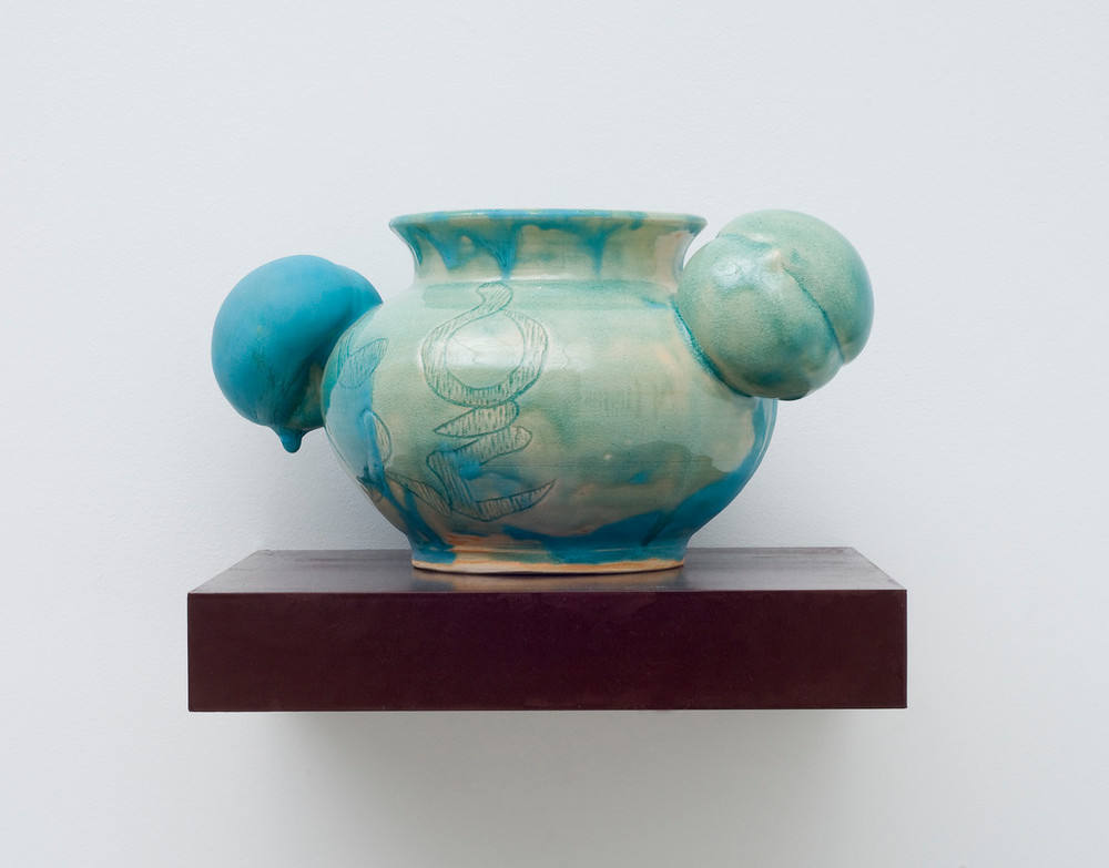 A round, ceramic vessel glazed unevenly in glossy celadon has a bulging body incised with abstract shapes. Matte turquoise glaze dribbling down from the rim obscures portions of the pale green and fully coats one of two peach-shaped orbs projecting from either side of the neck.