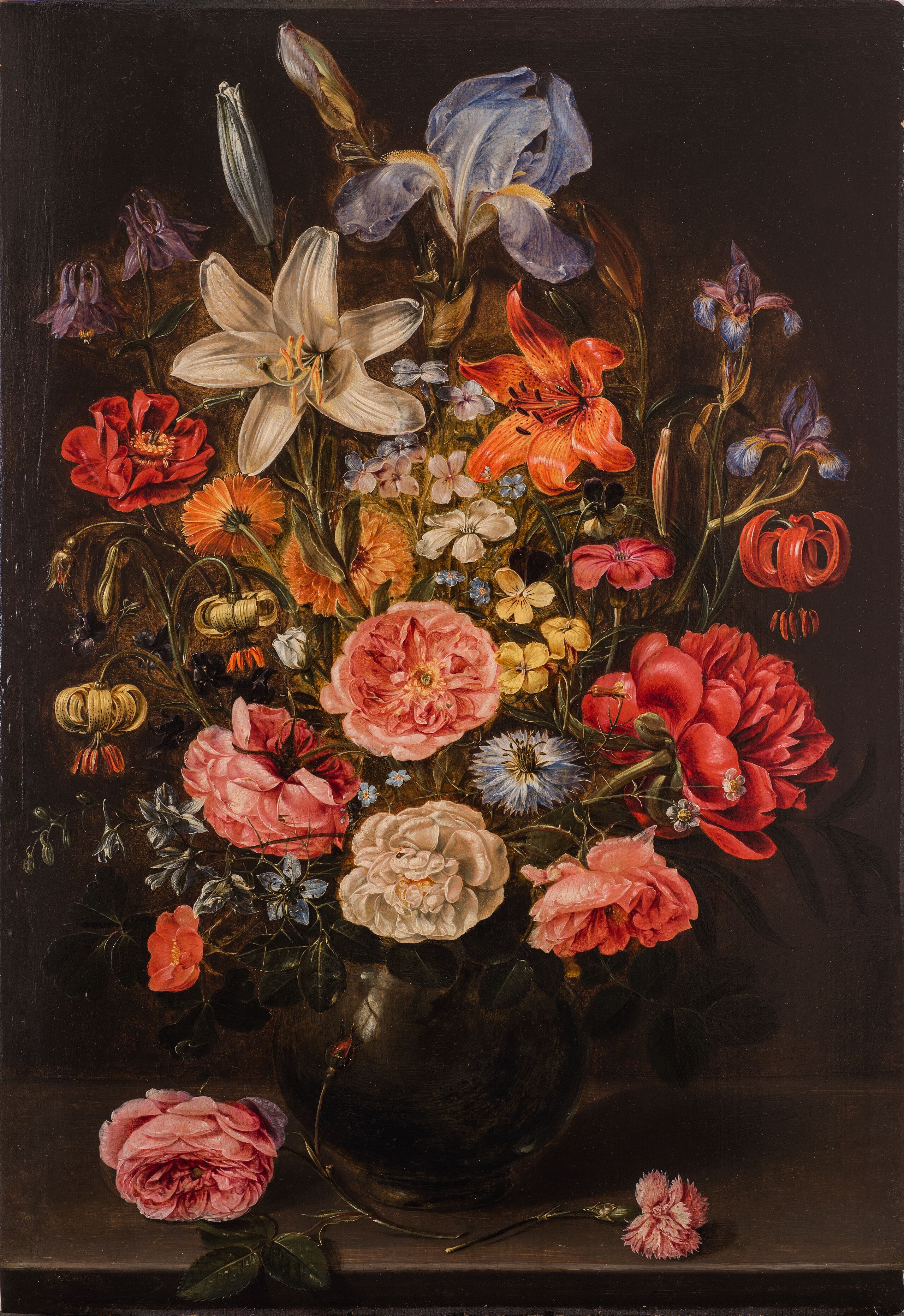 Realistic and detailed, a dramatic still-life painting features a large arrangement of varied, brightly colored flowers in a dark, round vase against a dark background. The vase sits on a stone ledge with two stray pink roses lying in the foreground.