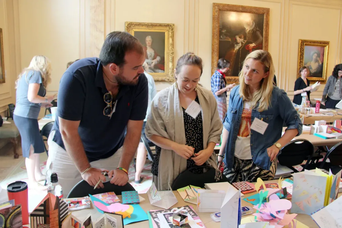 Three people with light skin tones stand in front of a table full of artists' books look at each other and chat.