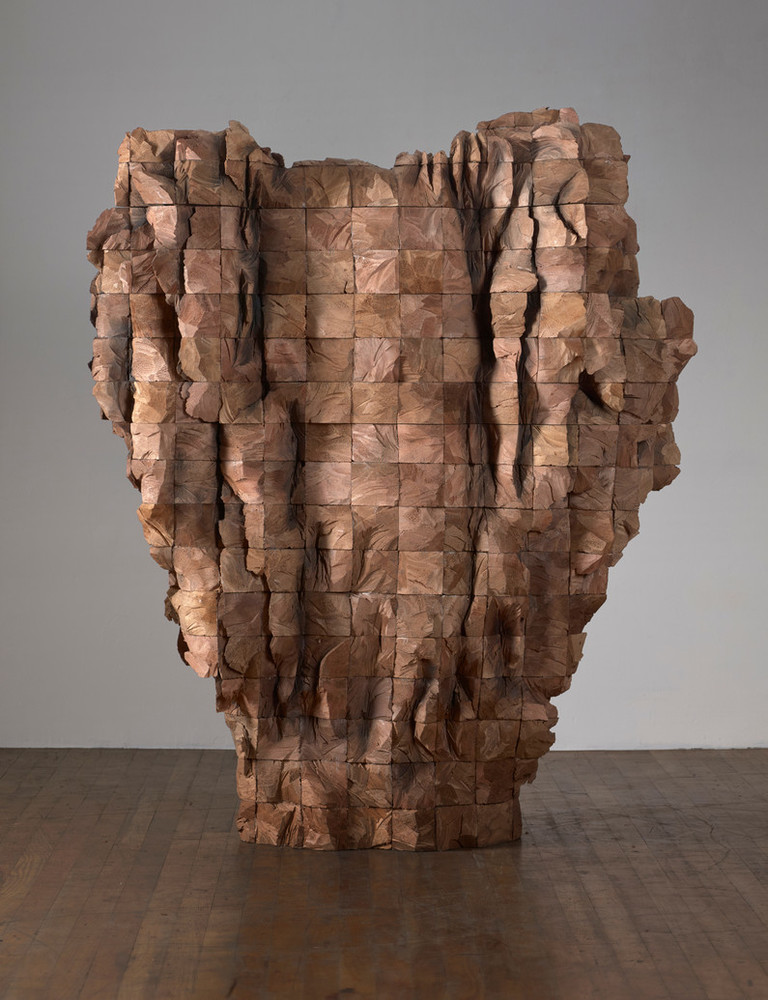 An abstract wooden sculpture composed of four-by-four beams of cedar stacked and roughly carved. Wide at the top, it tapers towards the bottom and sits directly on the floor.