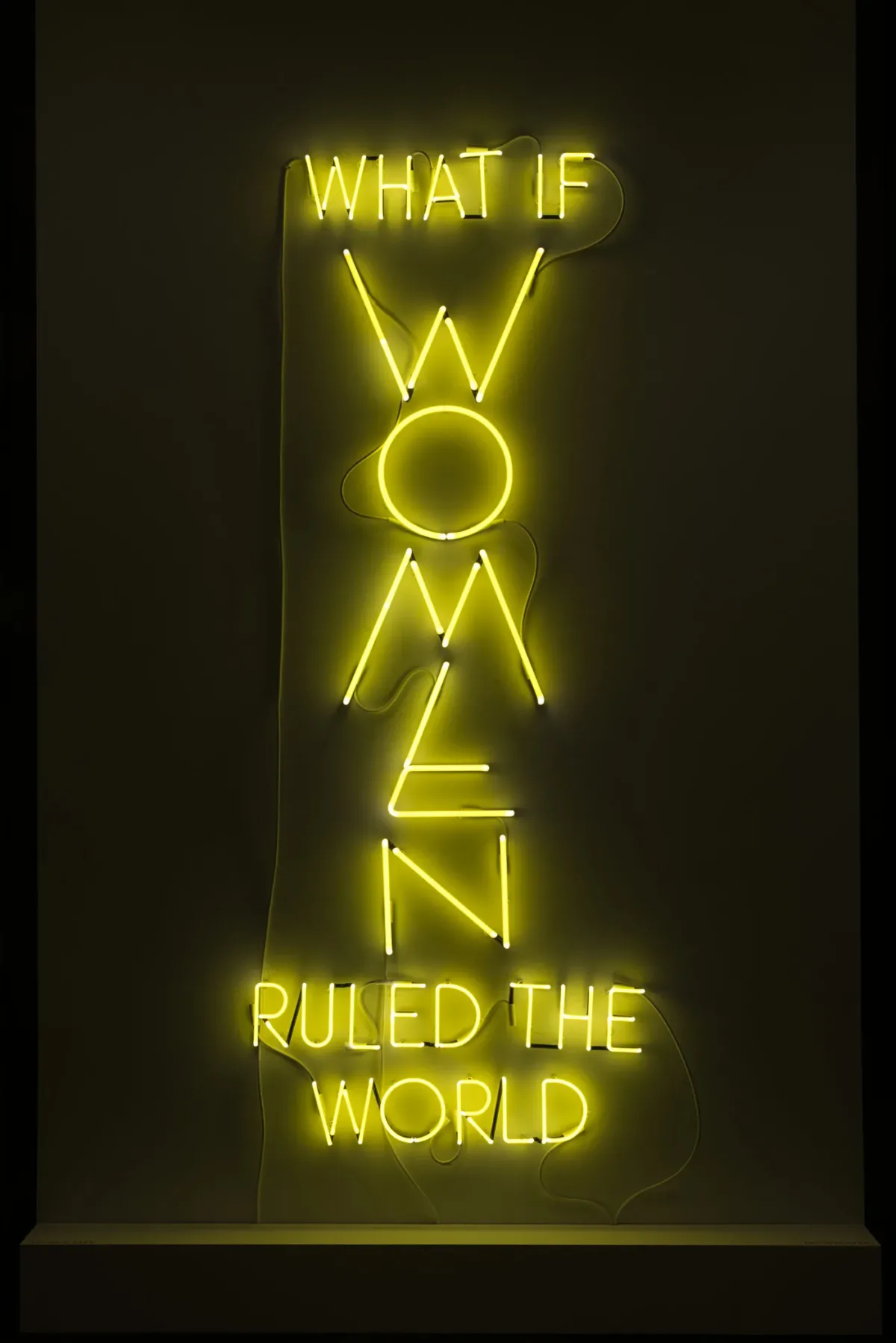 The phrase 'What if Women Ruled the World' is sculpted with neon tubes of bright yellow, in all capital letters. The letters of “WOMEN” are larger than the rest and are configured vertically.