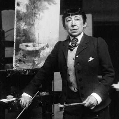 Black-and-white photograph of a light-skinned older woman with dark hair and bangs. She wears a dark suit with light-color vest, white collared shirt, and stripped necktie. She holds brushes in both hands and leans against an easel. Behind her is an unfinished painting.