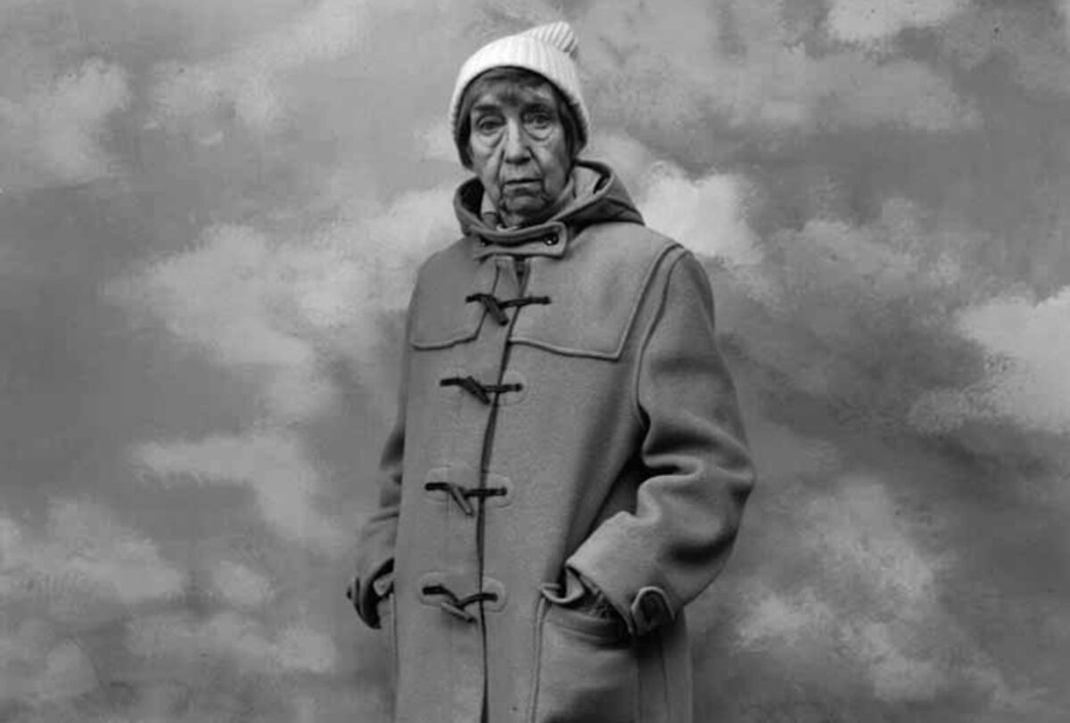 A black-and-white photograph of a light-skinned adult older woman wearing a long winter coat and white knitted cap. She has short hair and stands against a backdrop resembling a cloudy sky with her hands in her pockets.