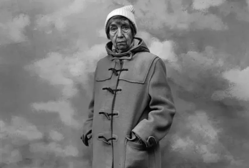 A black-and-white photograph of a light-skinned adult older woman wearing a long winter coat and white knitted cap. She has short hair and stands against a backdrop resembling a cloudy sky with her hands in her pockets.