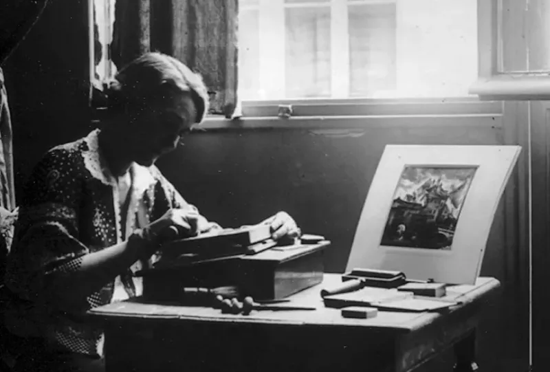 A black-and-white photograph of a woman seated at a table by a window. Her face, in profile, is in shadow. She looks down at a wood engraving. One hand steadies her woodblock while the other carves. Engraving and printing tools are scattered on the table, along with a small print.