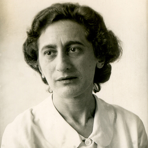 Black-and-white photograph of a light-skinned adult woman looking slightly to her right against a white background. Her dark, curly hair is styled neatly away from her face, and she wears a white collared button-up shirt.