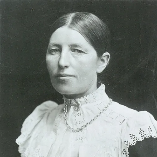 A black-and-white photograph of a light-skinned adult woman with her hair pulled back neatly. She smiles faintly and wears a lacy, high-collared, white dress with an anchor and chain brooch.