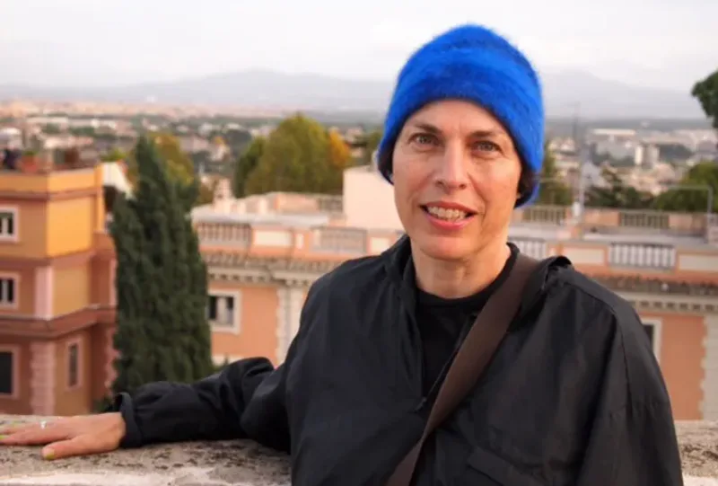 A light-skinned adult woman wearing a black zippered jacket, her pink lips parted in a smile. A royal blue, close-fitting hat covers her ears. Her right arm rests on a marble ledge. Behind her the rooftops of city buildings are visible.