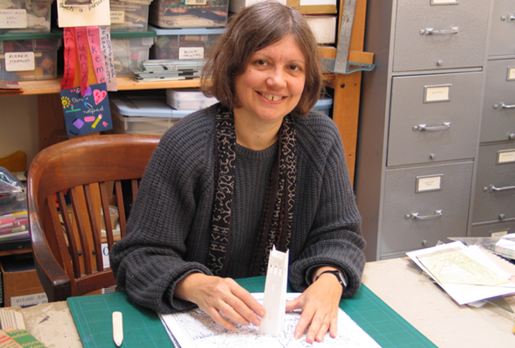 A light-skinned adult woman grins at the viewer, displaying a gap-toothed smile. She has chin-length gray hair and is working on a pop-up book depicting a tall white tower emerging from a white piece of paper on a cutting mat. Behind her are labeled bins of book art materials.